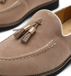 Close-up detail of taupe suede loafer showing gold metallic wrapped tassel and moc-toe stitching